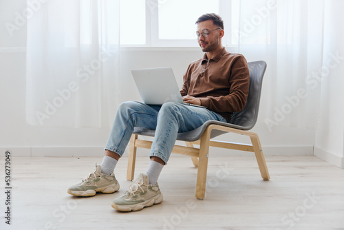Smiling cheerful happy young man guy in eyewear looks at screen laptop enjoys chatting with friends doing cool NFT project sitting on chair at home. Distance communication Remote Work New profession