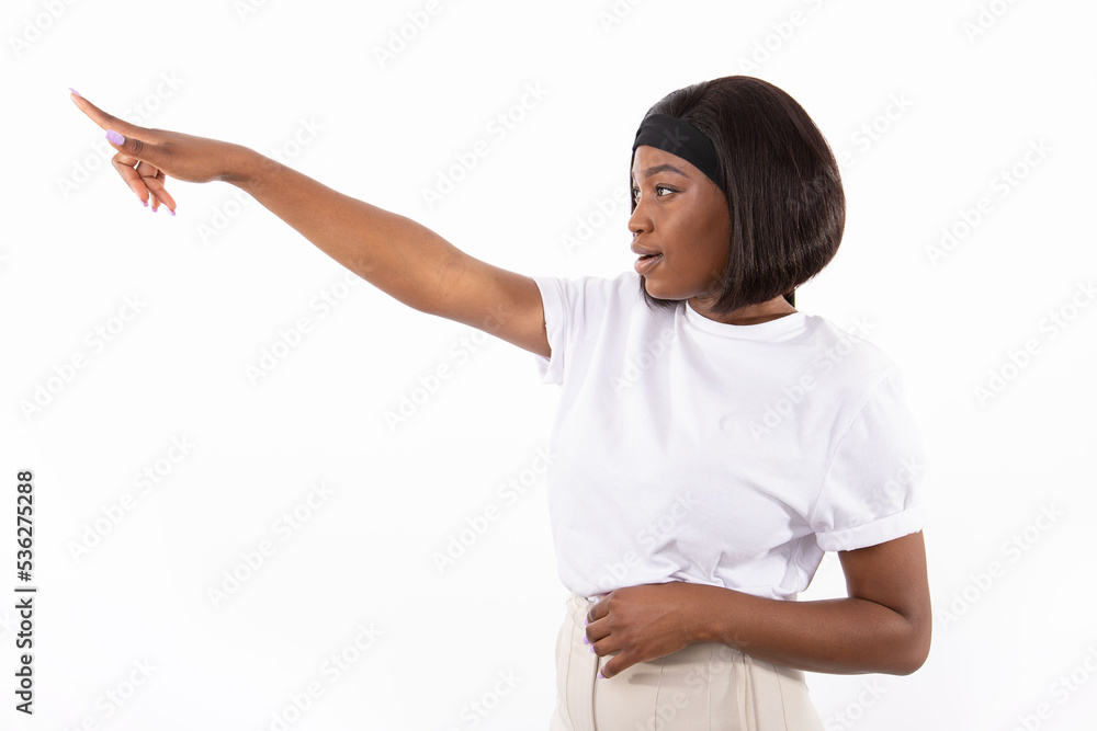 Portrait of happy young woman showing direction. African American lady wearing white T-shirt and headband looking away and pointing at something against white background. Advertising concept Portrait of happy young woman showing direction. African American lady wearing white T-shirt and headband looking away and pointing at something against white background. Advertising concept
