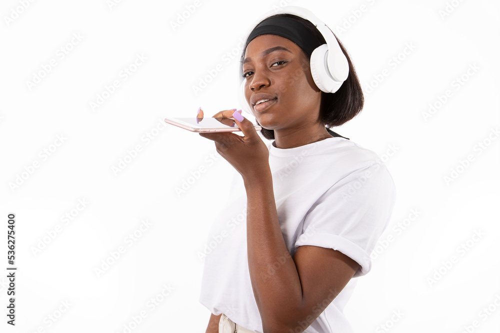 Portrait of happy young woman recording audio message in headphones. African American lady wearing white T-shirt and headband listening to music on mobile phone. Music and leisure concept Portrait of happy young woman recording audio message in headphones. African American lady wearing white T-shirt and headband listening to music on mobile phone. Music and leisure concept