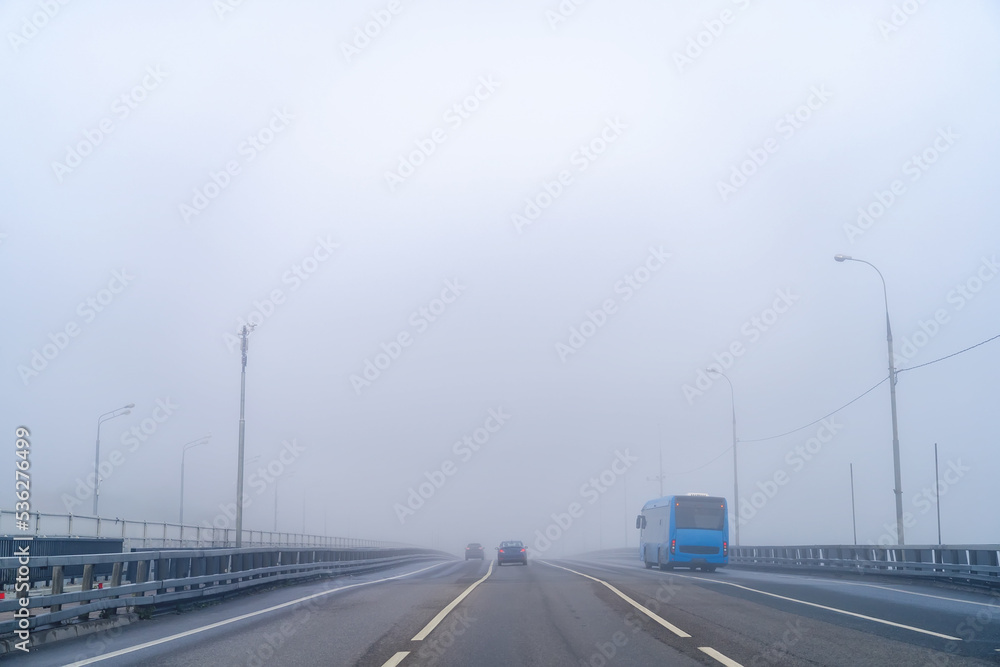 Blue bus and cars are driving along blue empty highway of city road with low visibility on warm foggy autumn morning. Blue urban electric bus in dense fog on autumn day.