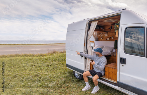 Hombre viajero caucásico, sentado en la camper, buscando ubicación en su teléfono móvil y señalando al paisaje.