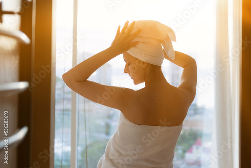 Female in white bathrobe and with towel at her head is standing on the living room after taking a shower in the early morning