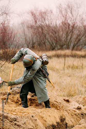 Re-enactor Dressed As German Wehrmacht Infantry Soldier In World War Ii With Shovel Digs Trench. Arrangement Of A Defensive Position. German Military Dress Of A German Soldier At World War II.