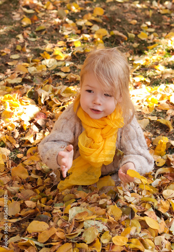 Cute little girl with long blonde hair sitting in big pile of yellow leaves. Child holding fallen yellow leaf in hand playing saying something