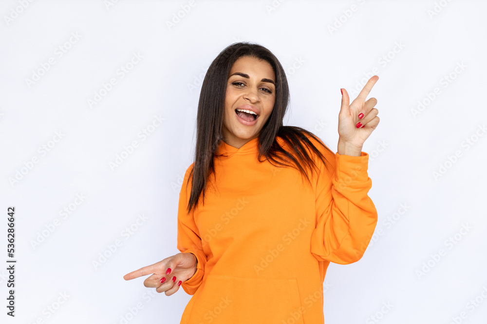 Portrait of excited Hispanic woman pointing in two ways. Happy young model with long hair in orange hoodie looking at camera, smiling while showing ads. Advertisement concept. Portrait of excited Hispanic woman pointing in two ways. Happy young model with long hair in orange hoodie looking at camera, smiling while showing ads. Advertisement concept.