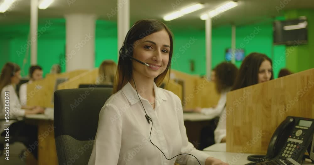 Portrait of young woman wearing headphone and working near computer at call center . Female emergency consultant , dispatcher or sales agent wears earphone , looks at camera and smiles . Slow motion Portrait of young woman wearing headphone and working near computer at call center . Female emergency consultant , dispatcher or sales agent wears earphone , looks at camera and smiles . Slow motion