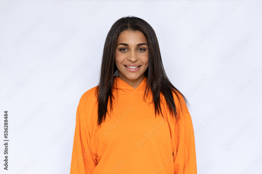 Portrait of happy Hispanic young woman smiling. Excited young female model with long hair in orange hoodie looking at camera, posing. Studio shot, happiness concept.