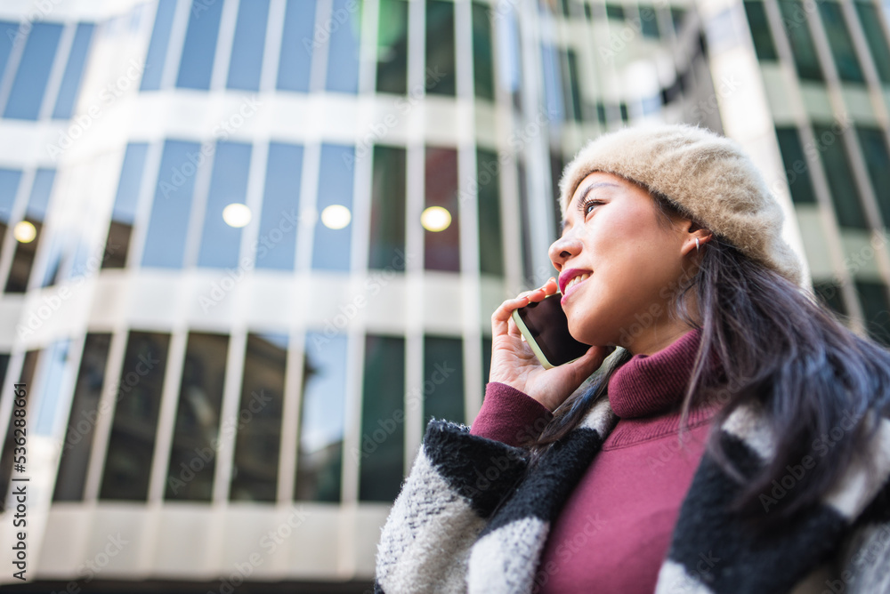 Fototapeta premium Side view of content Chinese female in outerwear and beret having phone conversation while standing near modern multistory building in city