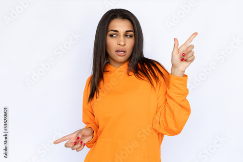 Portrait of doubtful Hispanic woman pointing in two ways. Thoughtful young model with long hair in orange hoodie looking away while showing ads. Advertisement concept. photo