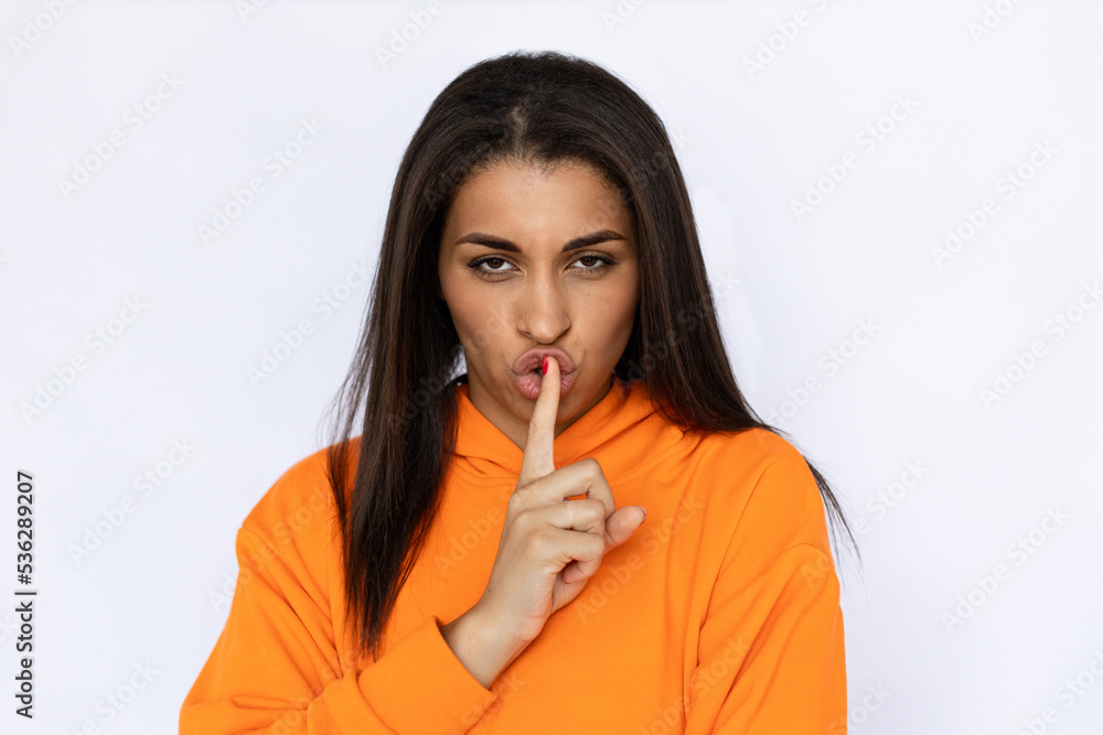 Portrait of frowned Hispanic woman shushing. Secretive young model with long hair in orange hoodie looking at camera with finger on lips, asking to be quiet. Secret, silence concept. Portrait of frowned Hispanic woman shushing. Secretive young model with long hair in orange hoodie looking at camera with finger on lips, asking to be quiet. Secret, silence concept.