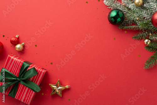 Christmas concept. Top view photo of pine branch decorated with green gold and red baubles star ornament big giftbox with ribbon bow and confetti on isolated red background with copyspace