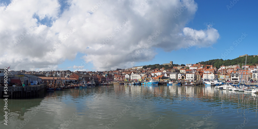 long panoramic view of the harbour in scarborough on a summer day with fishing boats reflected in the water and town buildings