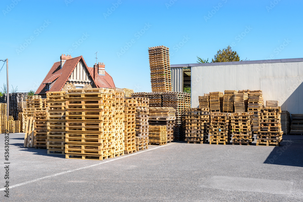 New and used wooden pallets stacked outdoors in the courtyard of a warehouse on a sunny day.
