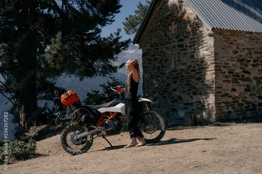 Female motorcyclist wearing dreadlocks standing near dirt motorcycle in long moto trip in mountains near a medieval stone building