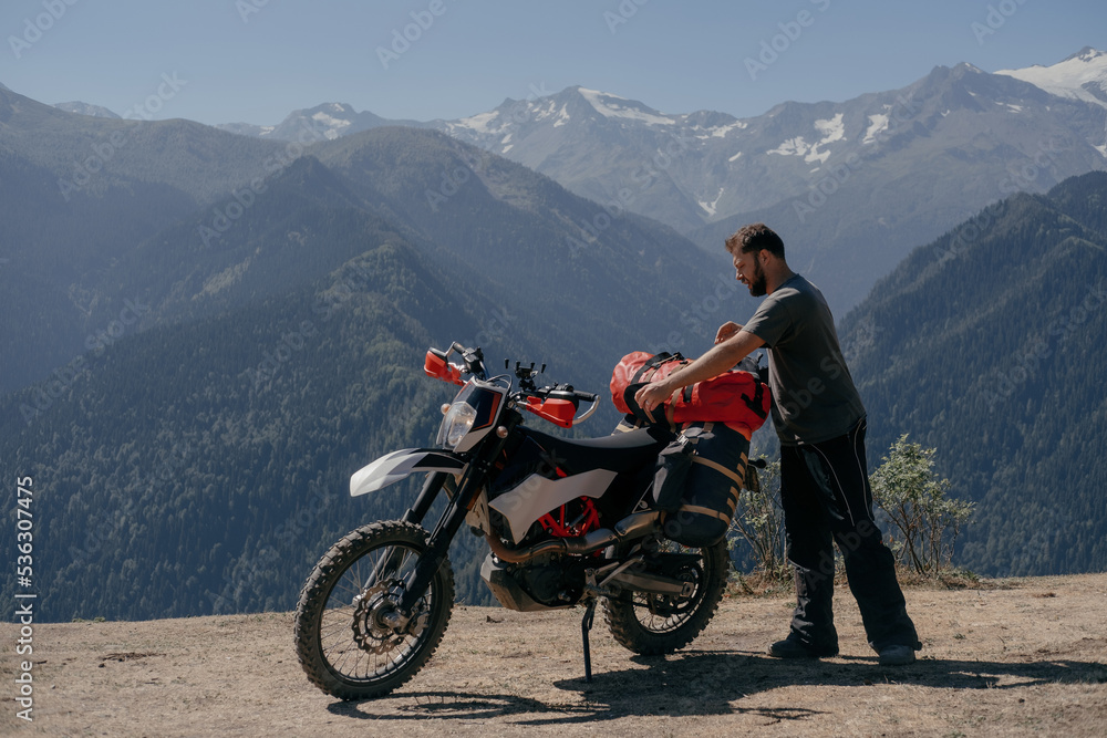 Biker man fastens dry bag on the trunk of dirt motorcycle in amazing mountainous summer landscape