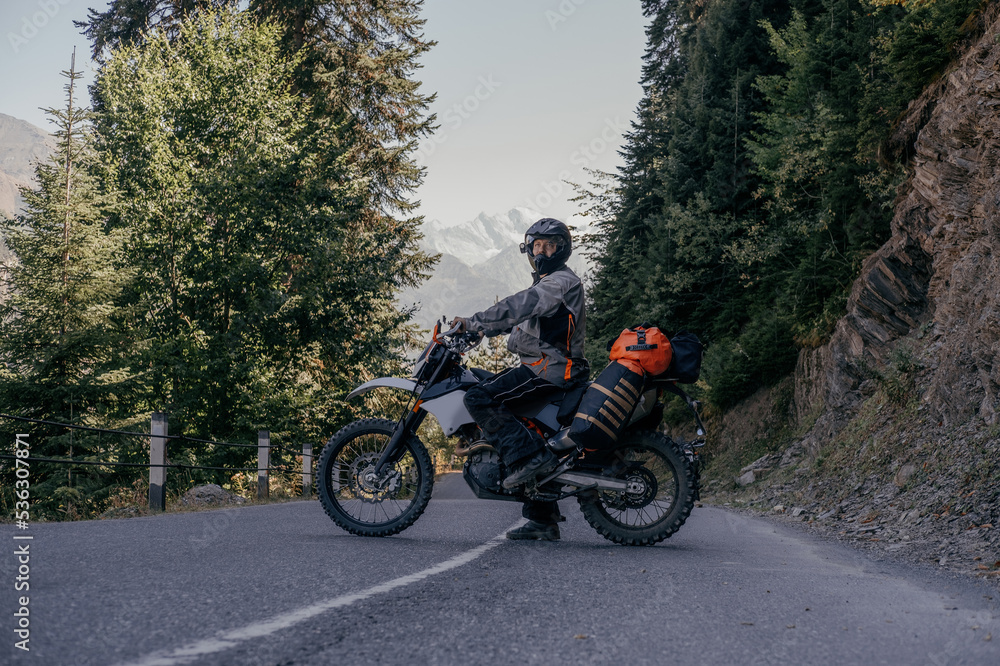 Biker man standing on dirt motorcycle loaded with big dry bag and wardrobe trunks on asphalt mountain road