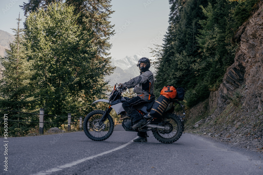 Biker man standing on dirt motorcycle loaded with big dry bag and wardrobe trunks on asphalt mountain road Biker man standing on dirt motorcycle loaded with big dry bag and wardrobe trunks on asphalt mountain road