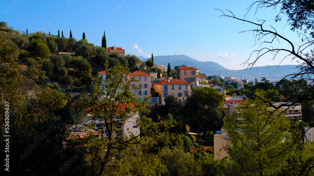 View of the village on the mountain in France, a panorama of the old town from the hill, a valley with tiny houses with orange roofs, green trees, and a blue sky, travel through Europe. View of the village on the mountain in France, a panorama of the old town from the hill, a valley with tiny houses with orange roofs, green trees, and a blue sky, travel through Europe.