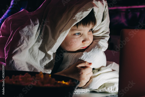 Happy kid son laughing eat popcorn remote control watching funny comedy tv show sitting on sofa having fun viewing video on laptop in evening at home. Teenager gen z boy under blanket at Movie night © Natallia
