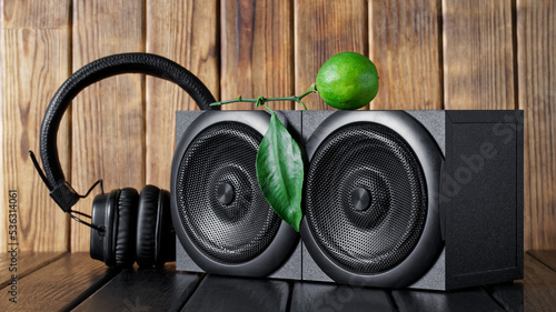 Two wooden audio speakers - acoustic system, installed in an interior made of natural pine planks next to wireless headphones and fresh lime green. Eco-style in music. Daylight.