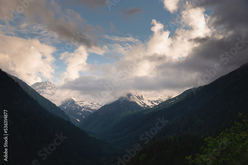 juicy green valley with rivers in grass and trees at the foot of snow-capped rocky mountains with waterfalls, voluminous fluffy thunderclouds through which sunlight breaks through © yanik88