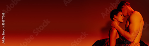 side view of man with muscular torso hugging sexy woman in bra on red background, banner.