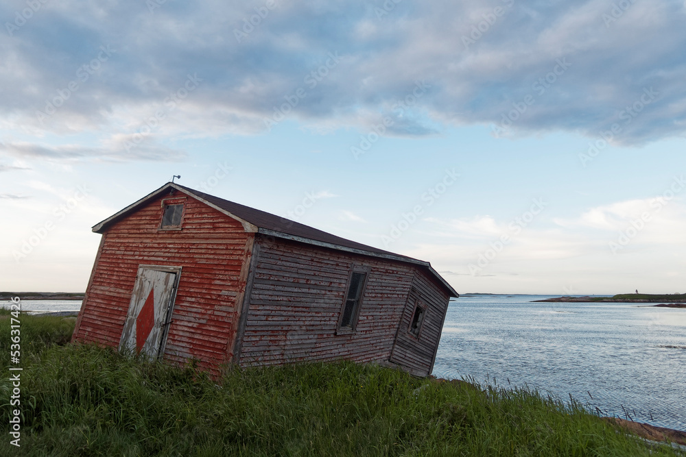 An old fishing shed near the ocean. The shed is red with a white and red door while showing extensive decay caused by abandonment.