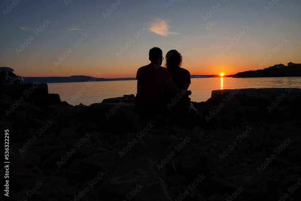 guy and girl, hugging romantic couple sitting on sea stone beach during sunset. soft light, mirroring and calm water. view from back on seashore on island, camping