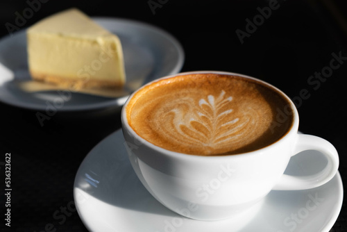 Latte art coffee in white cup whit cheese cake at back with dark background
