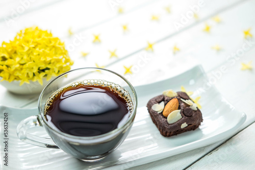Hot americano black coffee in clear cup on white plate with chocolate brownie cake beside place on old white table and beautiful flower around photo