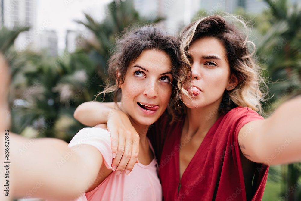 Carefree fair-haired lady making selfie during walk with friend. Blissful brunette girl embracing her sister on blur nature background.