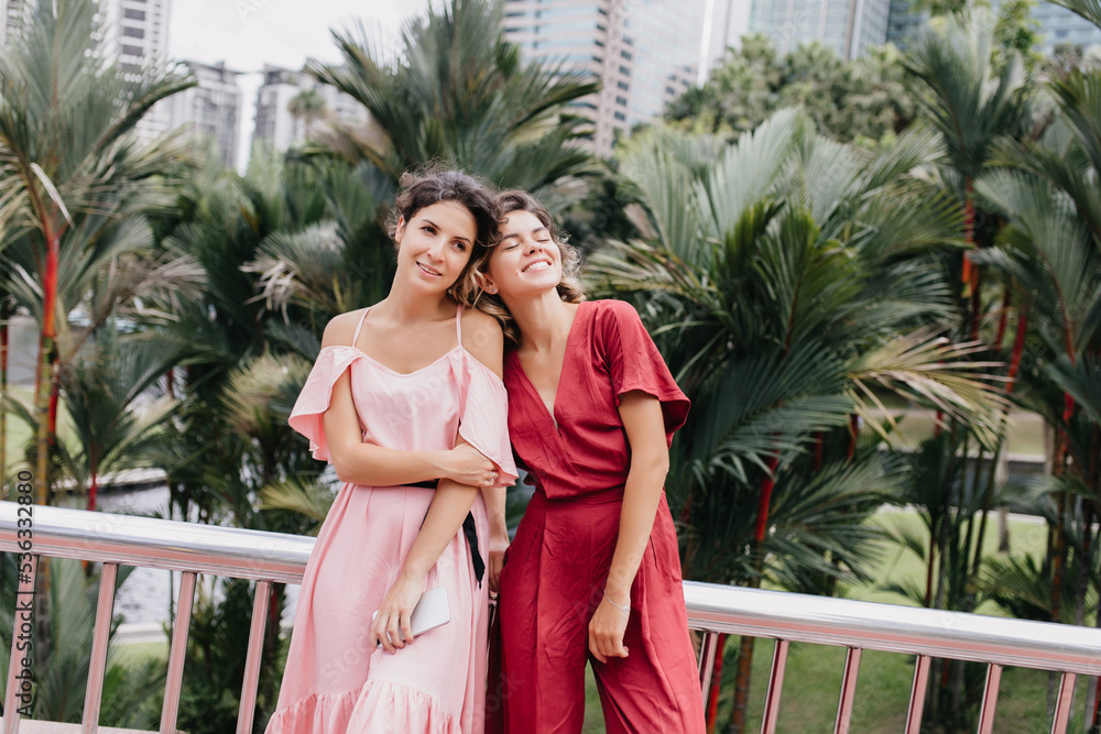 Amazing european woman in red dress standing on urban background beside friend. Joyful brunette girl expressing positive emotion during travel with sister. Amazing european woman in red dress standing on urban background beside friend. Joyful brunette girl expressing positive emotion during travel with sister.