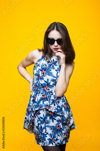 Portrait of a young woman in a dress and sunglasses on a yellow background.
