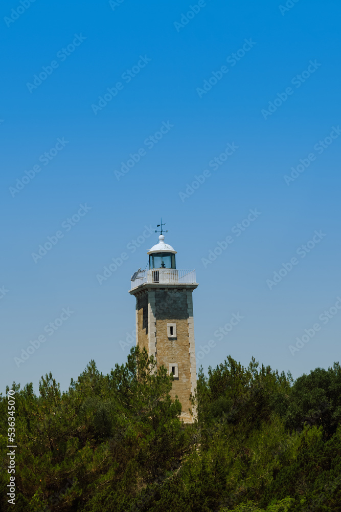 Day view of stone-built lighthouse surrounded by green trees against clear blue sky background. 