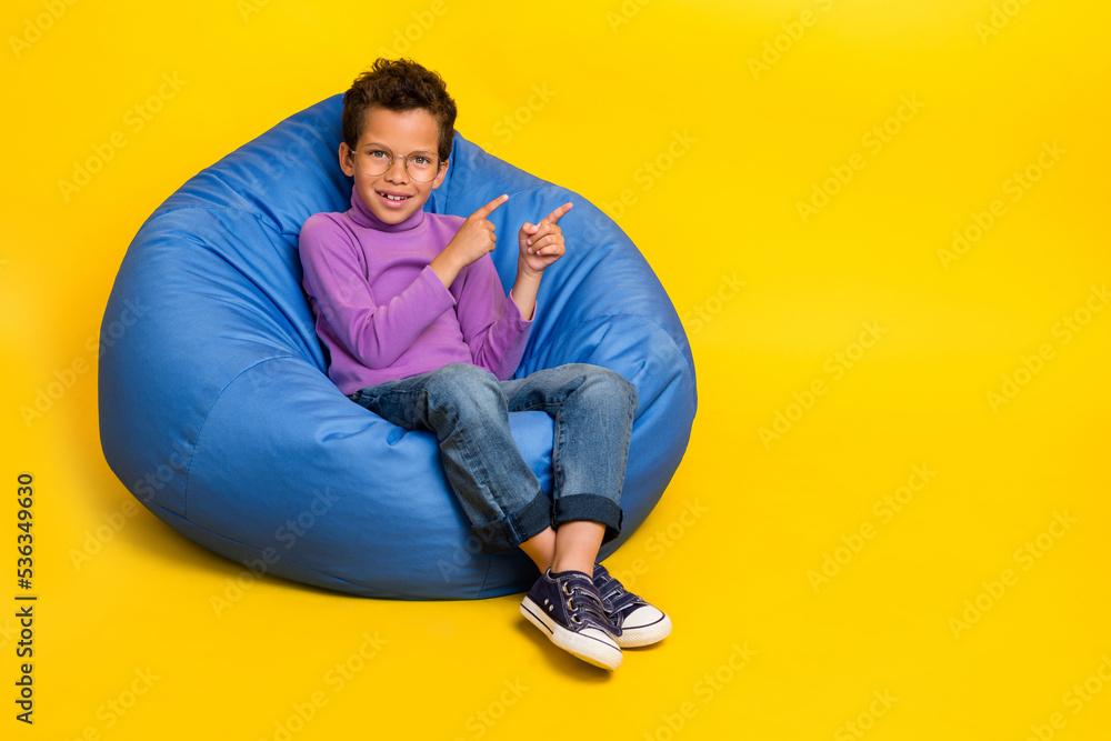 Full body photo of cheerful positive boy sit bag indicate fingers empty space isolated on yellow color background Full body photo of cheerful positive boy sit bag indicate fingers empty space isolated on yellow color background