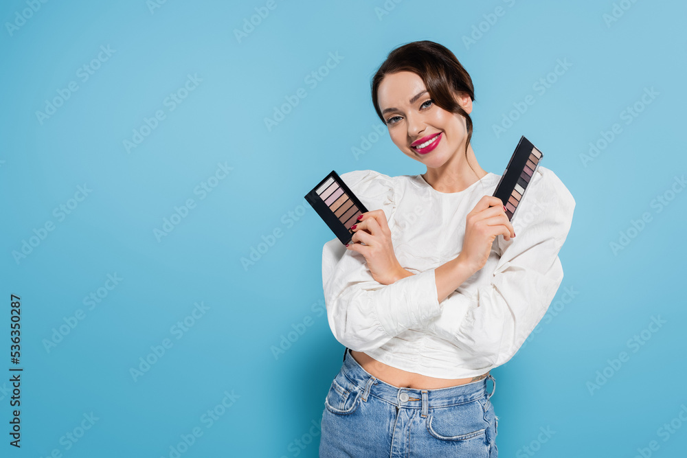 happy young woman in blouse holding eye shadow palettes on blue background. happy young woman in blouse holding eye shadow palettes on blue background.