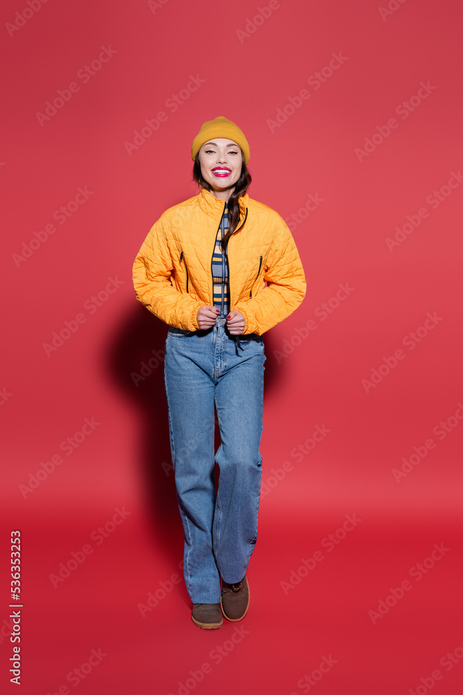 full length of happy young woman in beanie hat adjusting orange puffer jacket and smiling on red. full length of happy young woman in beanie hat adjusting orange puffer jacket and smiling on red.