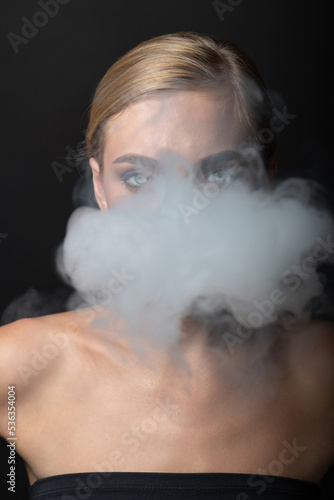 Fashion, style and make-up concept. Close-up studio portrait of beautiful blonde woman blowing smoke from mouth. Model looking to camera with green eyes and seductive look. Hair in camera focus