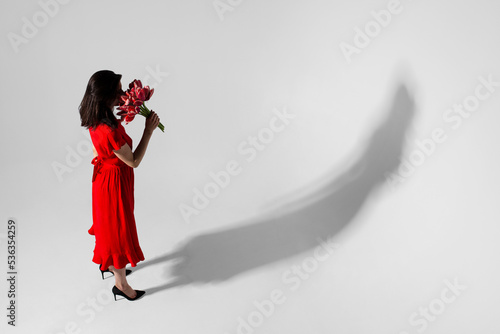 Full lengt view of a beautiful brunette young woman in red dress holding a bouquet of tulips and smells, isolated grey background.