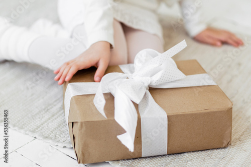 A girl in a white knitted dress sits on the floor and is about to open a gift in craft packaging with a white ribbon.