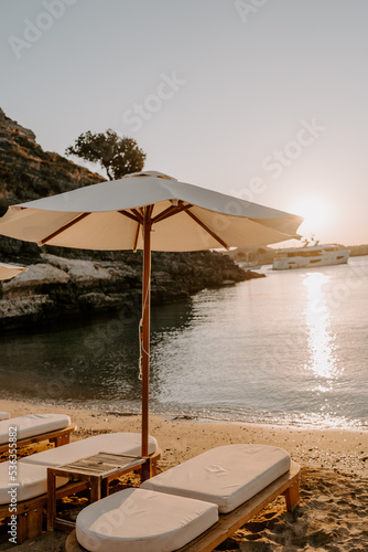 Fototapeta Naklejka Na Ścianę i Meble -  Tranquil beach scene with wooden sun loungers and white parasols lined along a pebbled shore. The turquoise sea, lush green mountains, and anchored yachts create a serene luxury escape in Türkiye