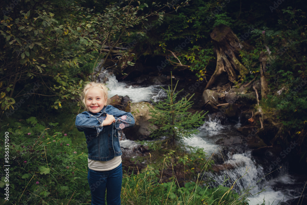 A little girl with blond hair, in jeans clothes, stands against the backdrop of a mountain river with stones and a forest, in summer. A little girl with blond hair, in jeans clothes, stands against the backdrop of a mountain river with stones and a forest, in summer.