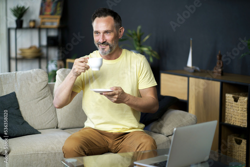 Enjoying coffee or tea middle aged business man make a break while working on laptop sitting on the sofa at home interior. Working distance mature IT man drinks coffee while working on computer