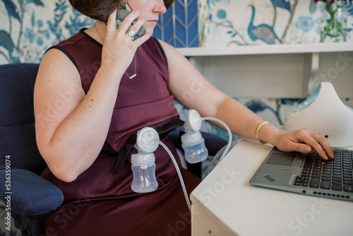 Woman on mobile phone and laptop while pumping at desk. photo