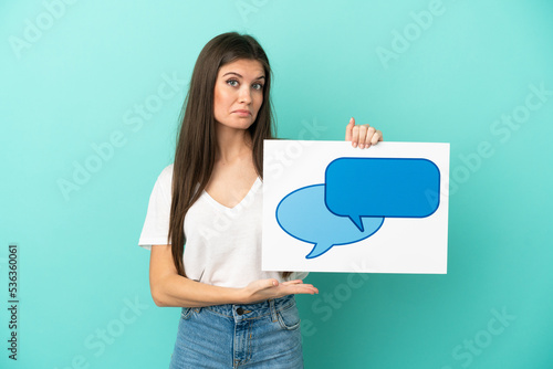 Young caucasian woman isolated on blue background holding a placard with speech bubble icon and pointing it