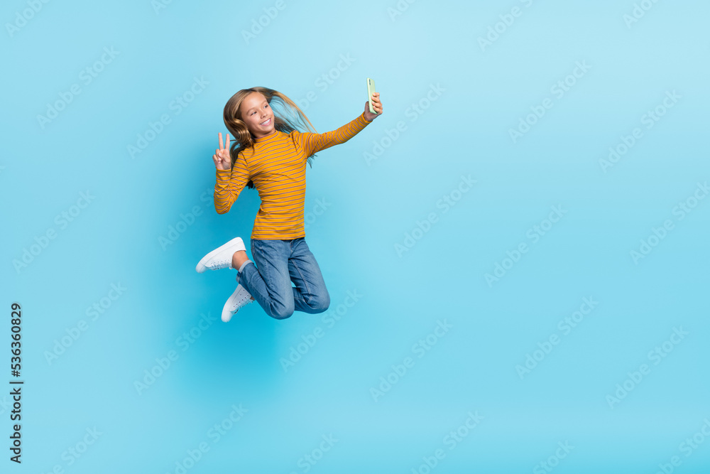 Full length portrait of energetic active girl jumping make selfie show v-sign isolated on blue color background Full length portrait of energetic active girl jumping make selfie show v-sign isolated on blue color background