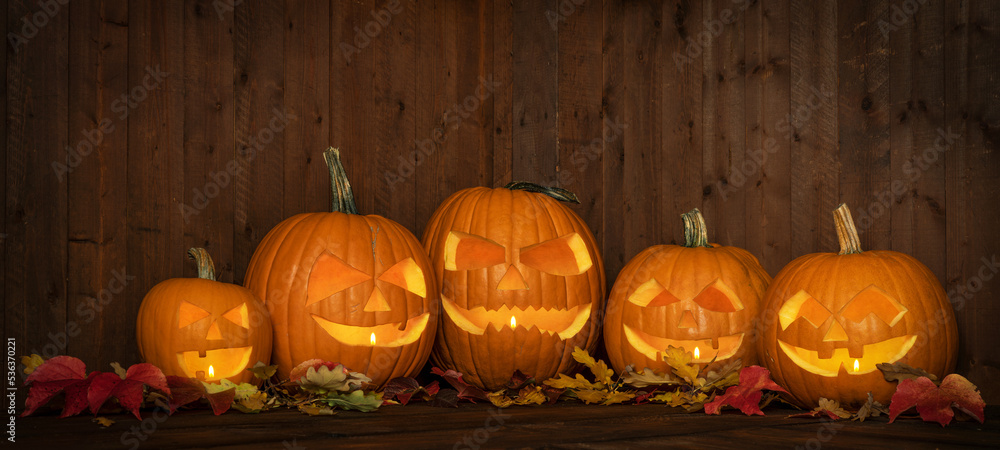 Obraz premium Scary Halloween celebration holiday party card background banner panorama - Many spooky carved glowing pumpkins, jack o lanterns and colorful fallen leaves on wooden table and wall
