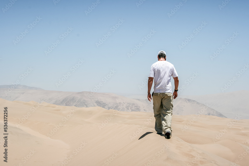 Person man with his back to the camera walking on the dunes in the desert at noon with a very strong sun.