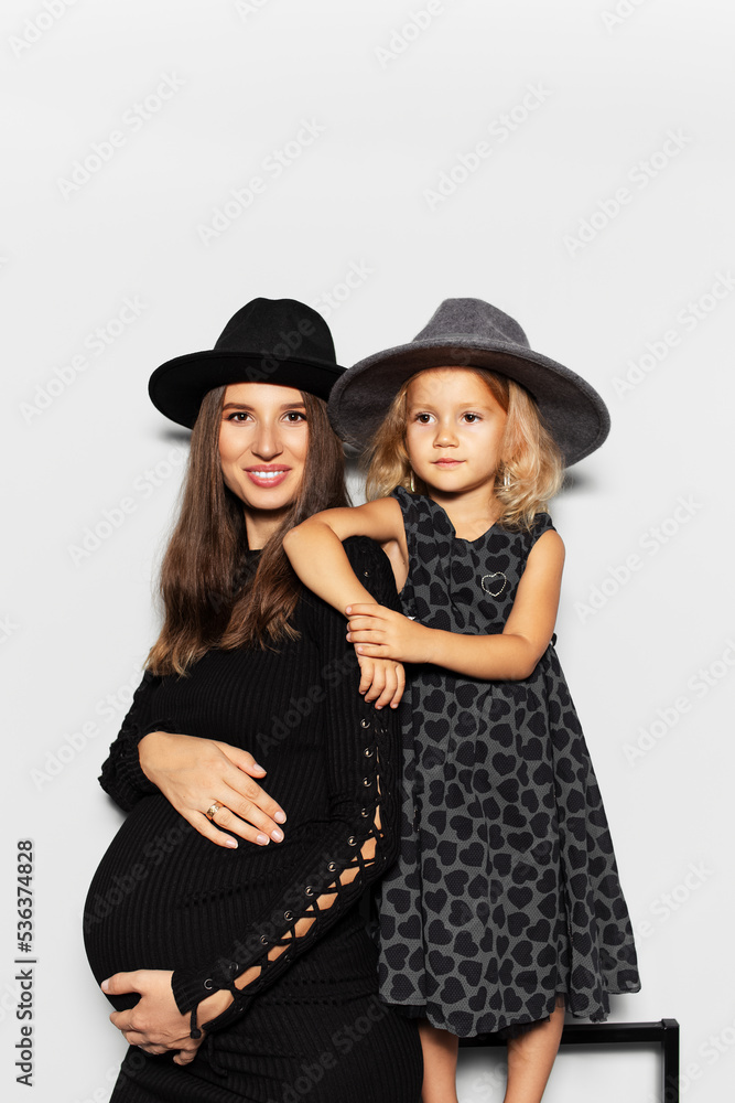 Studio portrait of pregnant smiling woman and child girl, mother and daughter, wearing hats on white background.