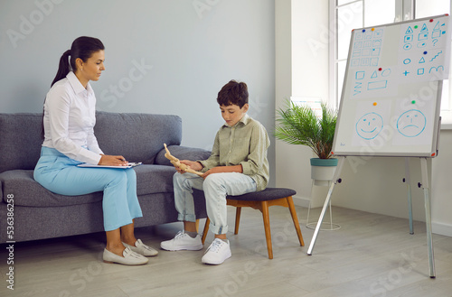 Professional psychologist, counselor or teacher working with little child. Woman with clipboard listening to school boy sitting on chair and talking about his problem. Psychology and education concept